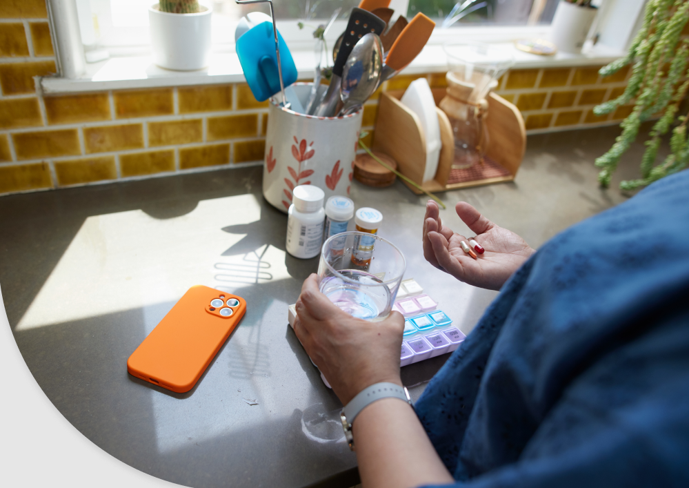 A woman at a kitchen counter with a glass of water, a weekly pill organiser and prescription bottles
