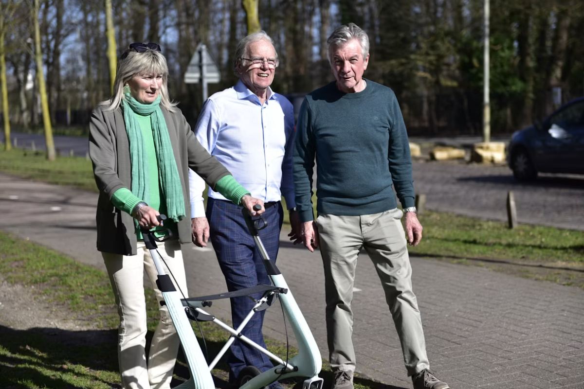 Three older adults walking together outdoors, one using a rollator