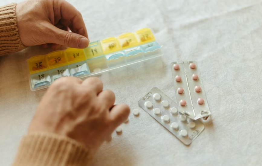 Hands filling a seven-day pill organiser at a table, blister packs beside it