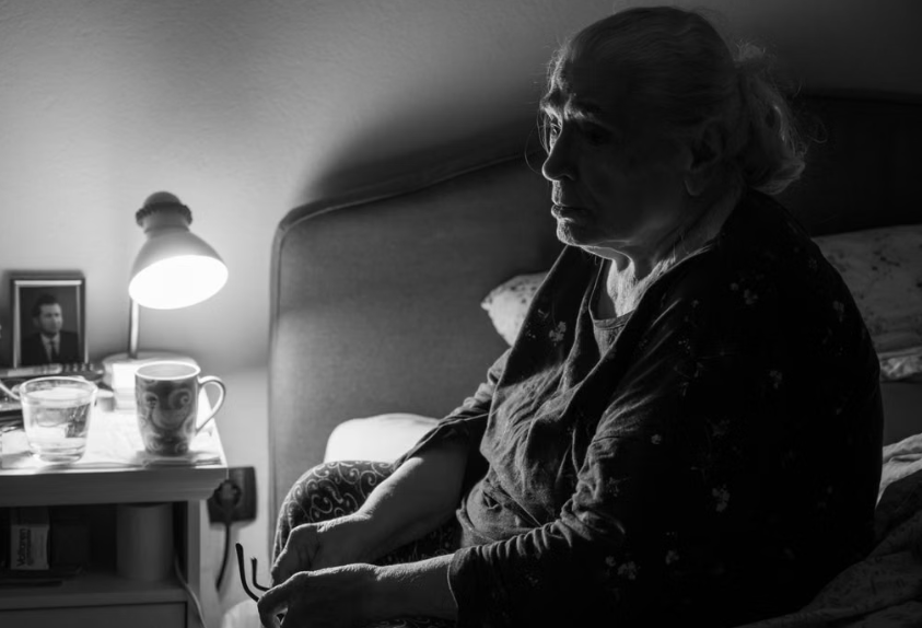An older woman sitting on the edge of her bed beside a lit lamp